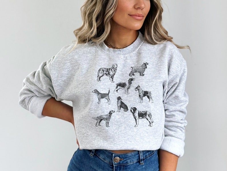 K&ouml;nnte beinhalten: Ein graues Sweatshirt mit einem Aufdruck verschiedener Hunderassen in Schwarzwei&szlig;. Die Hunde sind in einem Kreis auf der Vorderseite des Sweatshirts angeordnet.