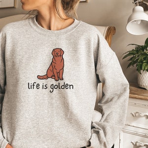 Op de afbeelding: Grijs sweatshirt met een cartoon golden retriever die zit en de tekst "life is golden" erop gedrukt.