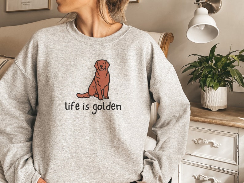 Op de afbeelding: Grijs sweatshirt met een cartoon golden retriever die zit en de tekst "life is golden" erop gedrukt.