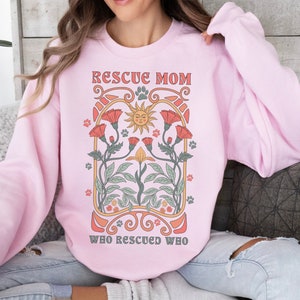 Puede incluir: Una sudadera rosa con el texto "Rescue Mom Who Rescued Who" en un dise&ntilde;o floral vintage. El dise&ntilde;o presenta un sol con cara, flores y huellas de patas.
