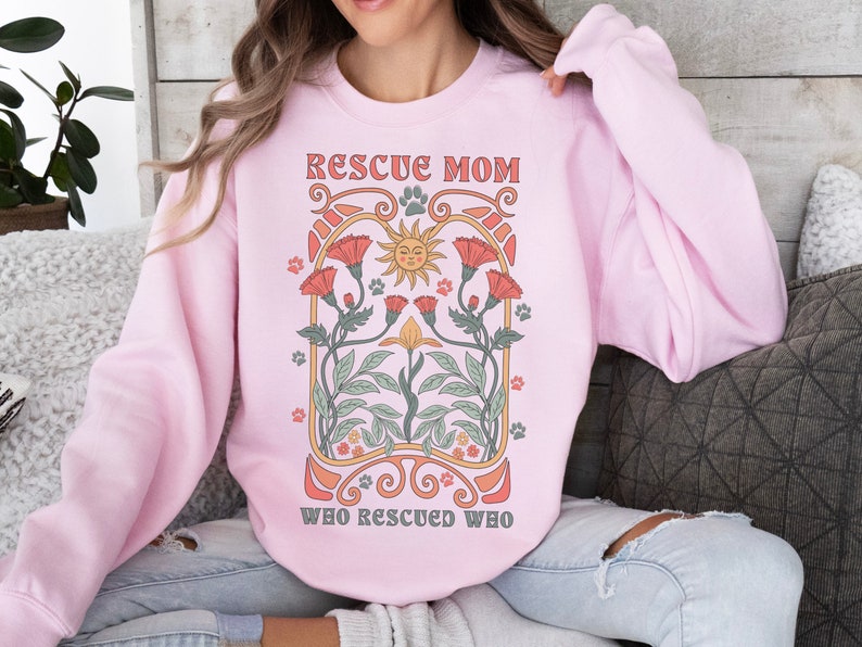 Puede incluir: Una sudadera rosa con el texto "Rescue Mom Who Rescued Who" en un dise&ntilde;o floral vintage. El dise&ntilde;o presenta un sol con cara, flores y huellas de patas.