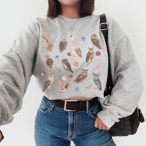 May include: A gray cropped sweatshirt with a print of owls, flowers, and butterflies. The sweatshirt has long sleeves and a crew neck.