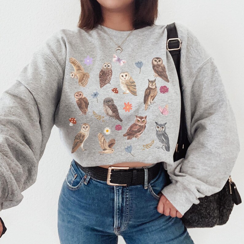 May include: A gray cropped sweatshirt with a print of owls, flowers, and butterflies. The sweatshirt has long sleeves and a crew neck.