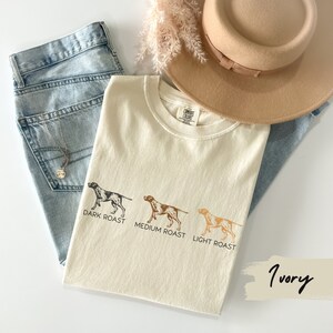 Op de afbeelding: Een wit T-shirt met een afbeelding van drie bruine honden die verschillende koffieroasters vertegenwoordigen: donker gebrande koffie, medium gebrande koffie en licht gebrande koffie. De tekst "DARK ROAST", "MEDIUM ROAST" en "LIGHT ROAST" staat onder elke hond gedrukt. Het T-shirt ligt op een blauwe denim achtergrond met een bruine hoed en een takje gedroogde bloemen.