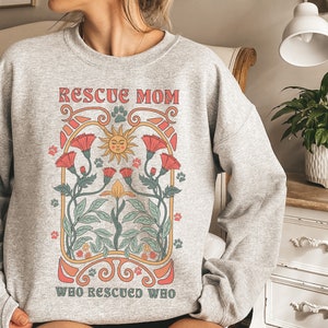 Puede incluir: Sudadera gris con un dise&ntilde;o gr&aacute;fico de un sol, flores y el texto "Rescue Mom Who Rescued Who".