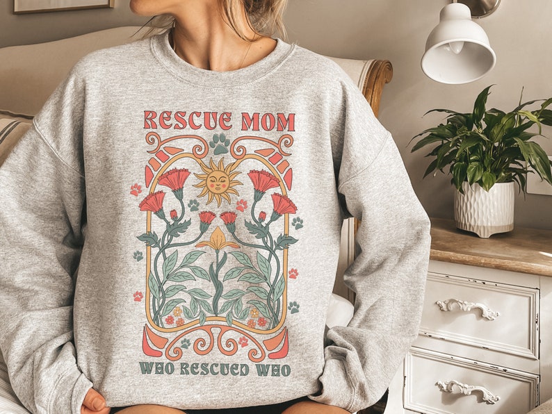 Puede incluir: Sudadera gris con un dise&ntilde;o gr&aacute;fico de un sol, flores y el texto "Rescue Mom Who Rescued Who".