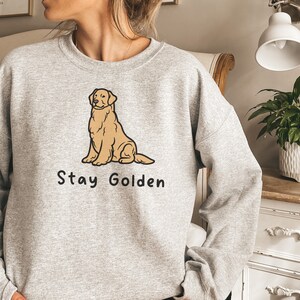 以下が含まれることがあります： 前面に卡通のゴールデンレトリバーが座っているイラストと「Stay Golden」という文字がプリントされた灰色のスウェットシャツです。
