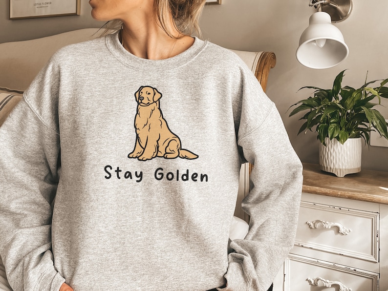 以下が含まれることがあります： 前面に卡通のゴールデンレトリバーが座っているイラストと「Stay Golden」という文字がプリントされた灰色のスウェットシャツです。
