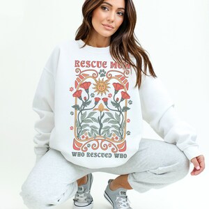 Puede incluir: Sudadera blanca con un dise&ntilde;o gr&aacute;fico que presenta un sol, flores y el texto "Rescue Mom Who Rescued Who".