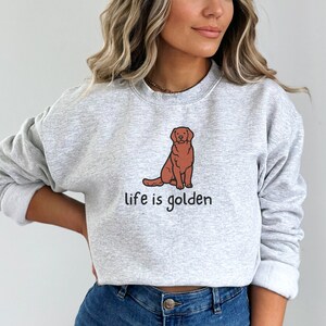Op de afbeelding: Een grijs sweatshirt met een bruine cartoon golden retriever en de tekst "life is golden" erop gedrukt.