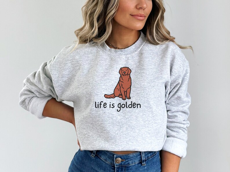 Op de afbeelding: Een grijs sweatshirt met een bruine cartoon golden retriever en de tekst "life is golden" erop gedrukt.