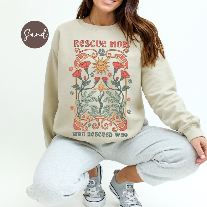 Puede incluir: Una sudadera beige con el texto "Rescue Mom Who Rescued Who" en una fuente retro. El dise&ntilde;o presenta un sol con una cara sonriente, flores y huellas de patas.