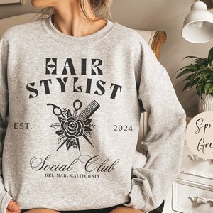 K&ouml;nnte beinhalten: Ein graues Sweatshirt mit dem Aufdruck "Hair Stylist Social Club" auf der Vorderseite. Der Text ist in schwarzer Schrift und von einer Schwarzwei&szlig;illustration einer Rose, einer Schere und eines Kamms umgeben. Der Text "Est. 2024" ist unter der Illustration gedruckt. Das Sweatshirt ist mit "Sport Grey" gekennzeichnet.