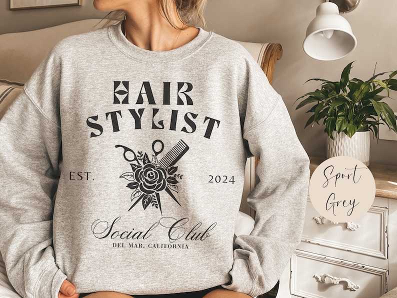 K&ouml;nnte beinhalten: Ein graues Sweatshirt mit dem Aufdruck "Hair Stylist Social Club" auf der Vorderseite. Der Text ist in schwarzer Schrift und von einer Schwarzwei&szlig;illustration einer Rose, einer Schere und eines Kamms umgeben. Der Text "Est. 2024" ist unter der Illustration gedruckt. Das Sweatshirt ist mit "Sport Grey" gekennzeichnet.