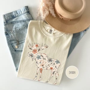 May include: Cream-coloured t-shirt with a floral moose design. The moose is filled with peach, blue, and cream-coloured flowers. The shirt is paired with light-wash denim jeans, a tan hat, and a gold necklace.