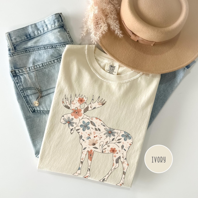 May include: Cream-coloured t-shirt with a floral moose design. The moose is filled with peach, blue, and cream-coloured flowers. The shirt is paired with light-wash denim jeans, a tan hat, and a gold necklace.
