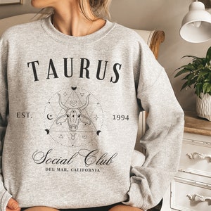 Puede incluir: Una sudadera gris con el texto "TAURUS EST. 1994 Social Club DEL MAR, CALIFORNIA" y un gr&aacute;fico en blanco y negro de una cabeza de toro dentro de un c&iacute;rculo con formas geom&eacute;tricas.
