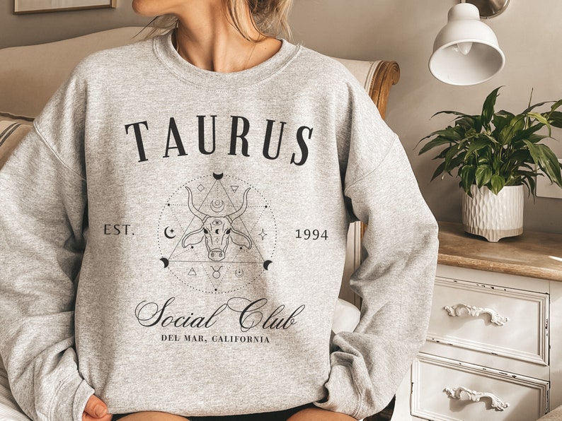 Puede incluir: Una sudadera gris con el texto "TAURUS EST. 1994 Social Club DEL MAR, CALIFORNIA" y un gr&aacute;fico en blanco y negro de una cabeza de toro dentro de un c&iacute;rculo con formas geom&eacute;tricas.