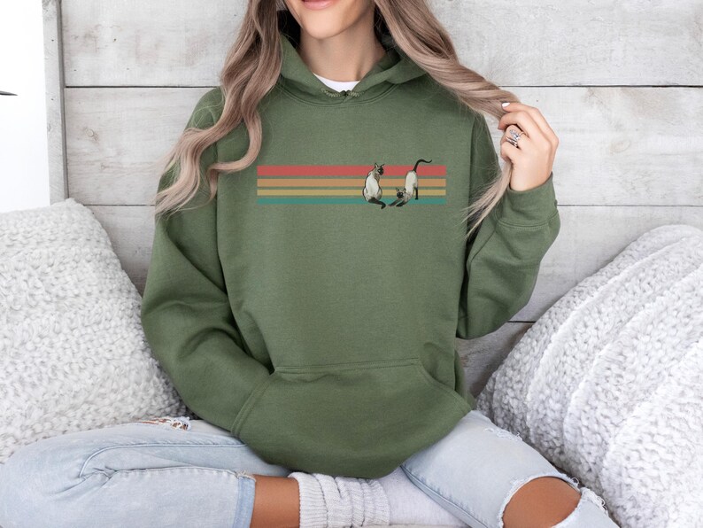 May include: Olive green hooded sweatshirt featuring a graphic of two Siamese cats beneath a retro-style horizontal stripe design in red, orange, yellow, and teal. The sweatshirt has a front pocket and a drawstring hood.