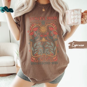 May include: A brown t-shirt with a graphic design featuring a sun, flowers, and the text "Rescue Mom Who Rescued Who".