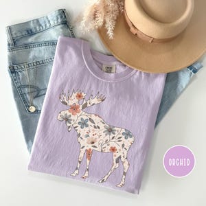 May include: A lilac t-shirt with a floral moose design. The moose is patterned with peach, blue, and cream flowers. The shirt is styled with light-wash jeans, a tan hat, and a necklace. The word "ORCHID" is in a purple circle.