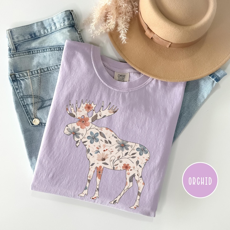 May include: A lilac t-shirt with a floral moose design. The moose is patterned with peach, blue, and cream flowers. The shirt is styled with light-wash jeans, a tan hat, and a necklace. The word "ORCHID" is in a purple circle.