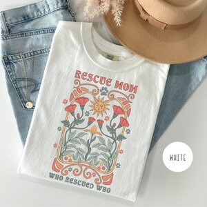 May include: White t-shirt with a graphic design of a sun, flowers, and paw prints. The text "Rescue Mom Who Rescued Who" is printed on the shirt.