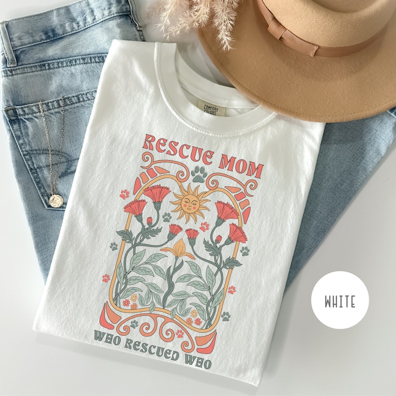 May include: White t-shirt with a graphic design of a sun, flowers, and paw prints. The text "Rescue Mom Who Rescued Who" is printed on the shirt.