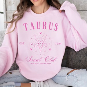 Puede incluir: Sudadera rosa con un dise&ntilde;o gr&aacute;fico que presenta la palabra "Taurus" y un toro estilizado con un c&iacute;rculo a su alrededor. El gr&aacute;fico tambi&eacute;n incluye el texto "Est. 1994" y "Social Club Del Mar, California".