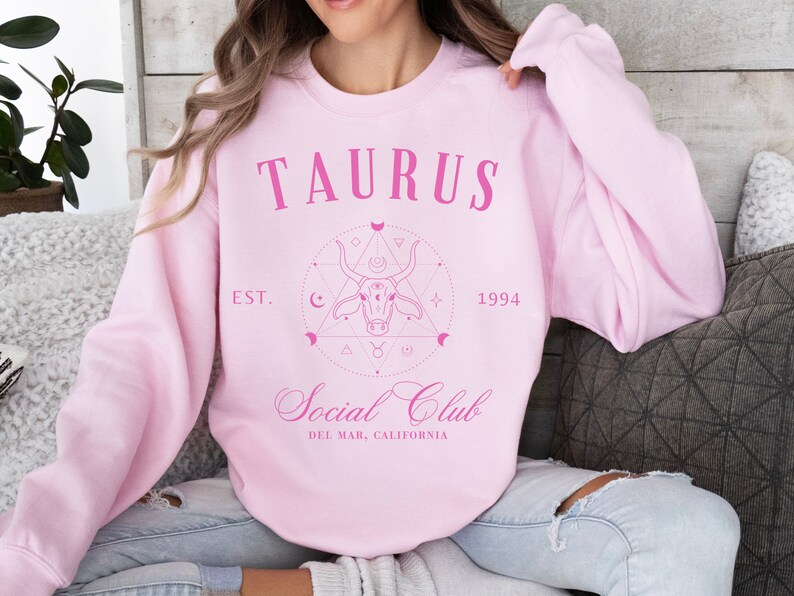 Puede incluir: Sudadera rosa con un dise&ntilde;o gr&aacute;fico que presenta la palabra "Taurus" y un toro estilizado con un c&iacute;rculo a su alrededor. El gr&aacute;fico tambi&eacute;n incluye el texto "Est. 1994" y "Social Club Del Mar, California".