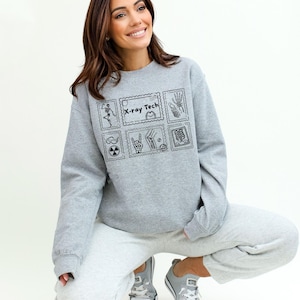 May include: A gray sweatshirt with a black graphic design featuring the text "X-ray Tech" and various medical-themed illustrations. The sweatshirt is paired with gray sweatpants and gray sneakers.