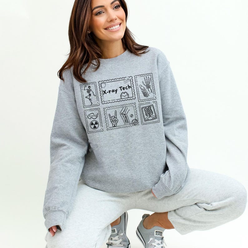May include: A gray sweatshirt with a black graphic design featuring the text "X-ray Tech" and various medical-themed illustrations. The sweatshirt is paired with gray sweatpants and gray sneakers.