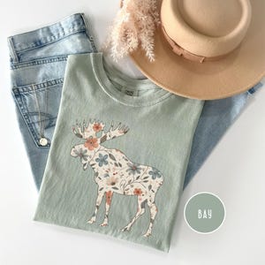 May include: A sage green t-shirt with a floral moose design, alongside light wash jeans, a tan hat, and a gold necklace. The moose is decorated with peach, blue, and cream flowers. A round sticker with the word "BAY" is also visible.