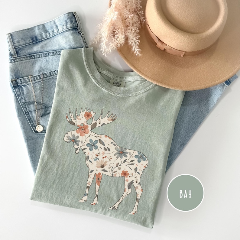 May include: A sage green t-shirt with a floral moose design, alongside light wash jeans, a tan hat, and a gold necklace. The moose is decorated with peach, blue, and cream flowers. A round sticker with the word "BAY" is also visible.