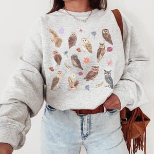 May include: A gray sweatshirt with a print of owls and flowers. The owls are in various poses and sizes, and the flowers are in shades of pink and purple.