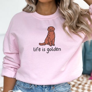 Op de afbeelding: Een roze sweatshirt met een cartoon golden retriever die zit en de tekst "life is golden" erop gedrukt.
