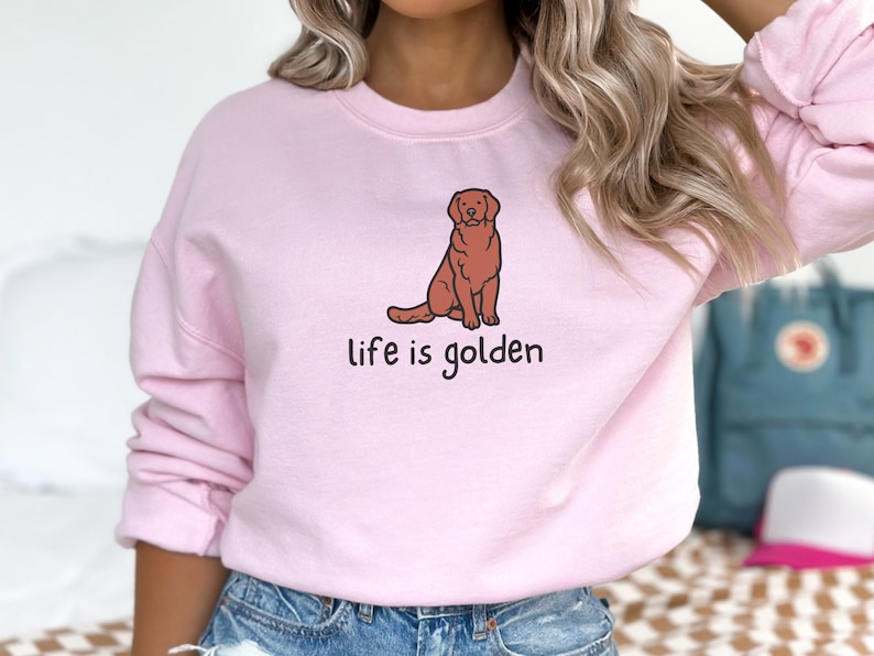 Op de afbeelding: Een roze sweatshirt met een cartoon golden retriever die zit en de tekst "life is golden" erop gedrukt.