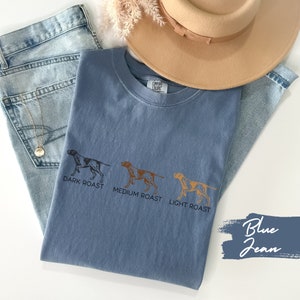 Op de afbeelding: Een jeansblauw T-shirt met drie illustraties van een hond die verschillende koffieroasters voorstellen: donker gebrande koffie, medium gebrande koffie en licht gebrande koffie. De tekst "Dark Roast", "Medium Roast" en "Light Roast" staat onder elke hond gedrukt. Het T-shirt is gevouwen en ligt op een wit oppervlak met een blauwe spijkerbroek en een bruine hoed.