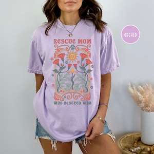 May include: A lavender t-shirt with a graphic design of a sun, flowers, and paw prints. The text on the shirt reads "Rescue Mom Who Rescued Who".