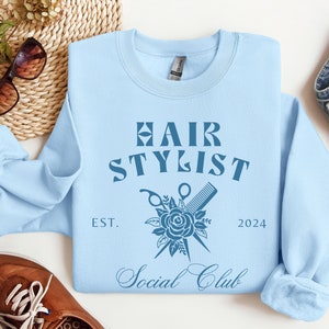 K&ouml;nnte beinhalten: Hellblauer Pullover mit dem Text "Hair Stylist Social Club" in Blau, mit einem stilisierten Rosen- und Scherendesign. Der Text "Est. 2024" befindet sich unter dem Design.