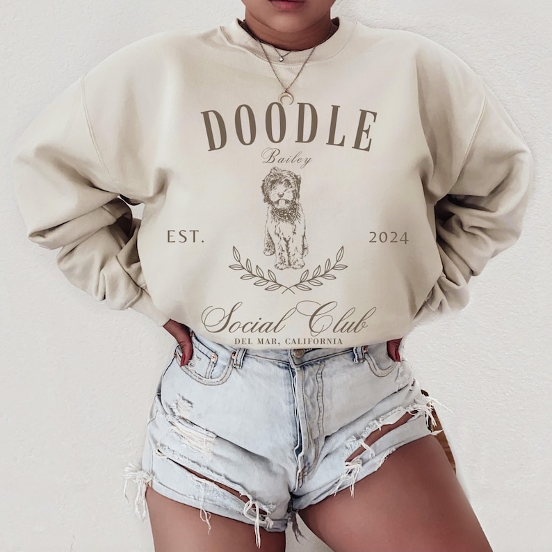 Puede incluir: Una sudadera beige con el texto "Doodle Bailey Est. 2024 Social Club Del Mar, California" y un garabato de un perro.