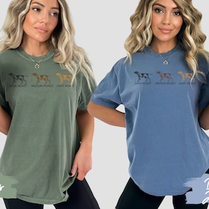 Op de afbeelding: Twee vrouwen dragen T-shirts met een afbeelding van drie honden en de tekst "DARK ROAST MEDIUM ROAST LIGHT ROAST". De vrouw links draagt een groen shirt en de vrouw rechts draagt een blauw shirt.