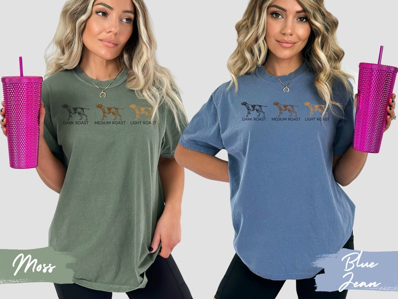 Op de afbeelding: Twee vrouwen dragen T-shirts met een afbeelding van drie honden en de tekst "DARK ROAST MEDIUM ROAST LIGHT ROAST". De vrouw links draagt een groen shirt en de vrouw rechts draagt een blauw shirt.