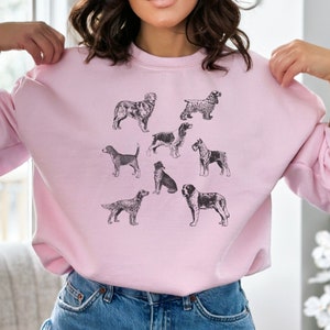 K&ouml;nnte beinhalten: Ein rosa Sweatshirt mit einer Schwarzwei&szlig;illustration verschiedener Hunderassen. Die Hunde sind in verschiedenen Posen dargestellt, darunter sitzend, stehend und laufend.
