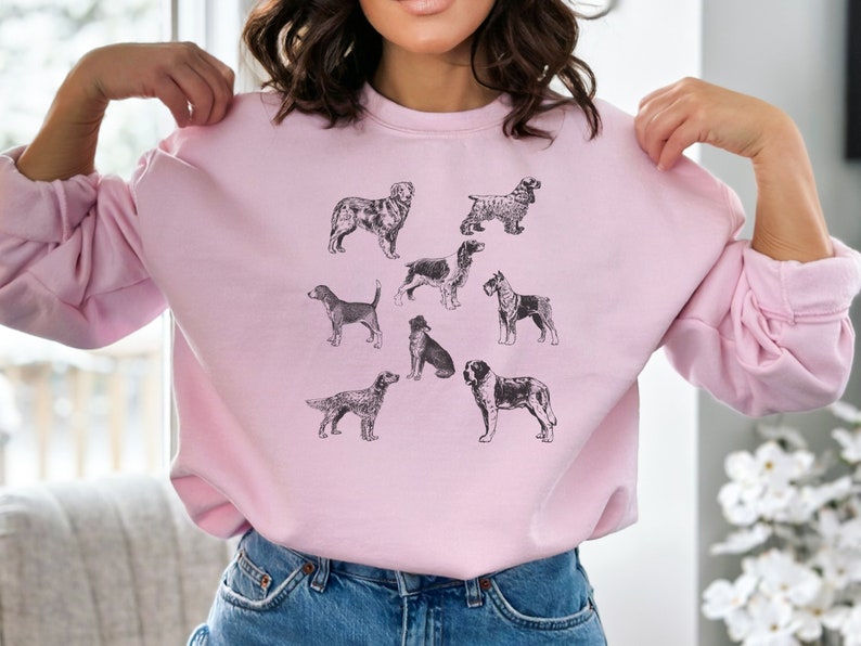 K&ouml;nnte beinhalten: Ein rosa Sweatshirt mit einer Schwarzwei&szlig;illustration verschiedener Hunderassen. Die Hunde sind in verschiedenen Posen dargestellt, darunter sitzend, stehend und laufend.