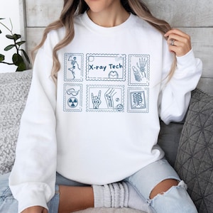 May include: White sweatshirt featuring blue illustrations of X-ray-related images, including a skeleton, hand, and the text "X-ray Tech." The design is arranged in a grid pattern, resembling postage stamps. The sweatshirt is a casual, comfortable garment.