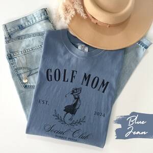 K&ouml;nnte beinhalten: Ein blaues Jeans-T-Shirt mit dem Text "GOLF MOM" und einer Grafik einer Frau, die Golf spielt. Der Text "EST. 2024" und "Social Club TORREY PINES" befindet sich ebenfalls auf dem Shirt.
