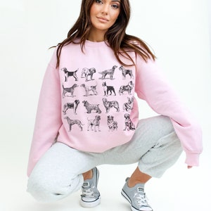 May include: A pink sweatshirt with a graphic print of various dog breeds in black and white. The model is wearing the sweatshirt with grey sweatpants and white sneakers.