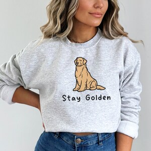 以下が含まれることがあります： 前面に卡通ゴールデンレトリバーが座っているイラストと「Stay Golden」という文字がプリントされたグレーのトレーナーです。