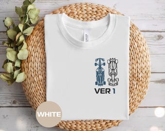 May include: White t-shirt with a crew neck. The shirt features two blue race car illustrations and the text "VER 1" printed below. The shirt is displayed on a woven, circular surface.